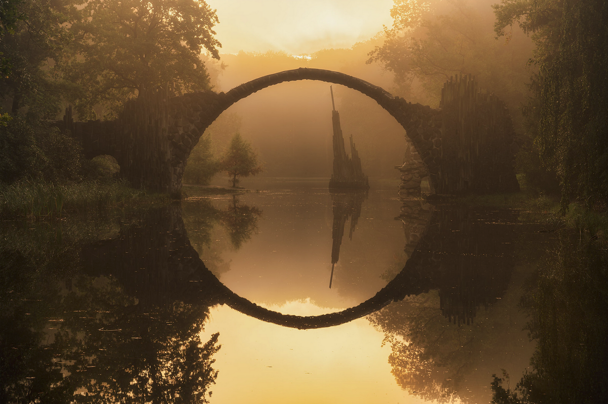 The arch of the Devil's Bridge in Germany during sunrise, drawing a circle on the horizon thanks to the lake underneath it.